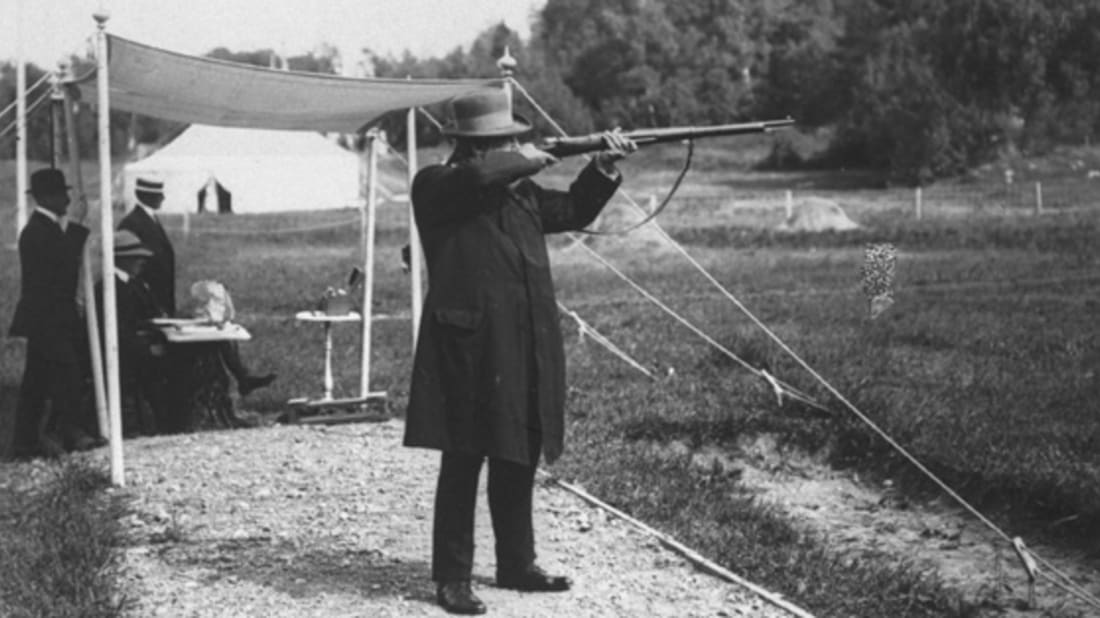 Tir aux pigeons vivants aux Jeux Olympiques de Paris 1900, un tireur vise un pigeon lâché depuis une trappe, sous une tente d’époque